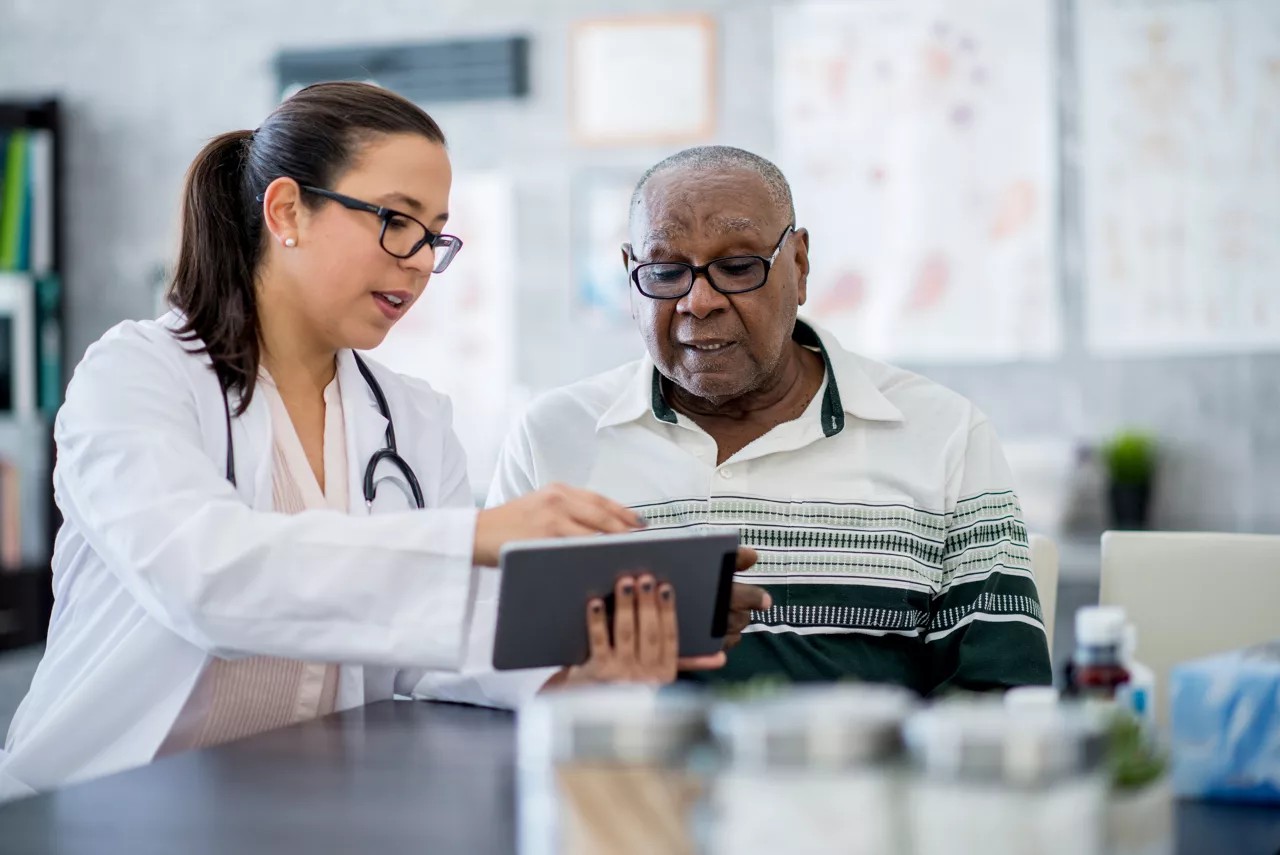 A senior man of African descent is indoors in a hospital room, looking at a tablet with his doctor. The female doctor is explaining his medication schedule, utilizing the tablet for visual aid.